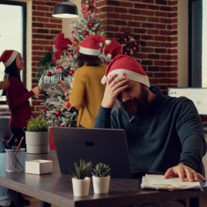 Employee being frustrated by christmas holiday festivity in startup office, trying to work on laptop during xmas eve time. Stressed man being disturbed and interrupted by noisy people.